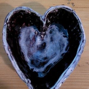 May include: A heart-shaped ceramic bowl featuring a glossy, dark purple interior glaze with a lighter, mottled lavender and white center. The exterior rim is thick and textured with a pale, uneven finish that contrasts with the dark basin.