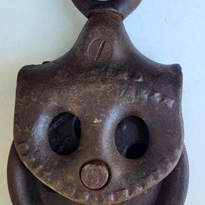 Puede incluir: Polea de metal oxidado de &eacute;poca, con un gran ojal en la parte superior y una rueda en la parte inferior. La polea tiene dos grandes agujeros redondos y un perno central. El metal tiene un aspecto envejecido y desgastado.