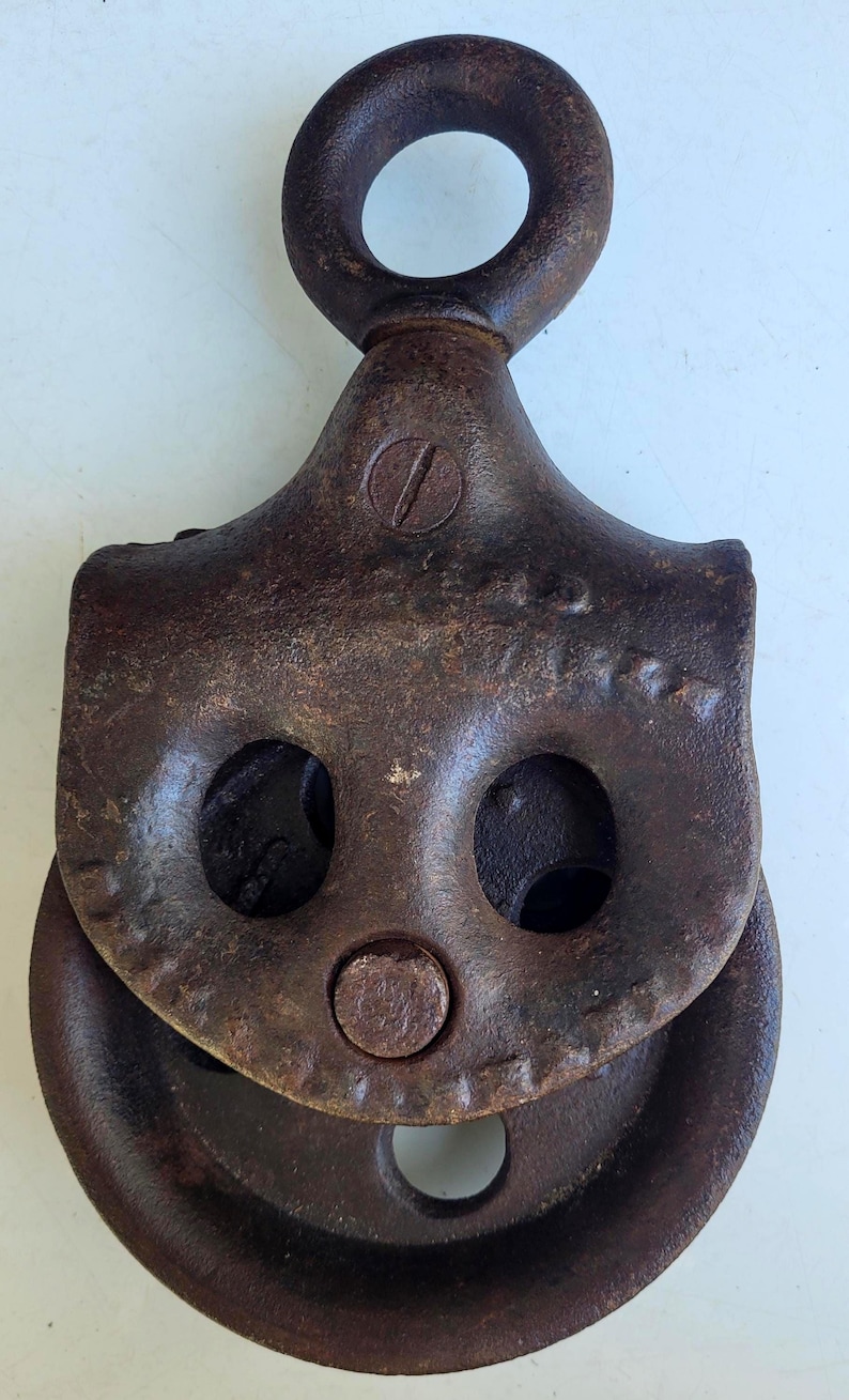 Puede incluir: Polea de metal oxidado de &eacute;poca, con un gran ojal en la parte superior y una rueda en la parte inferior. La polea tiene dos grandes agujeros redondos y un perno central. El metal tiene un aspecto envejecido y desgastado.