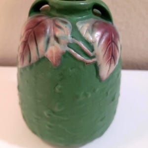 May include: A matte green ceramic vase featuring a textured surface and two small handles at the top. The upper portion of the vase is decorated with two prominent, raised leaf motifs in shades of deep red and muted purple, adding a distinct visual element to the cylindrical body of the vessel.