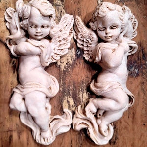 May include: Two off-white cherub figurines with detailed wings and draped fabric accents. Each cherub has curly hair and a neutral expression, featuring a matte finish with subtle brown antiquing in the crevices of the sculpted forms.