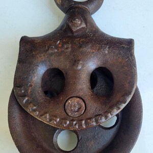 Puede incluir: Una polea de metal oxidado y vintage con un lazo superior circular y una rueda acanalada. La polea tiene dos grandes agujeros en forma de ojo y un remache central. El metal tiene un aspecto desgastado y envejecido, lo que sugiere que es una antig&uuml;edad.