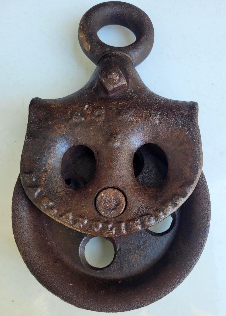 Puede incluir: Una polea de metal oxidado y vintage con un lazo superior circular y una rueda acanalada. La polea tiene dos grandes agujeros en forma de ojo y un remache central. El metal tiene un aspecto desgastado y envejecido, lo que sugiere que es una antig&uuml;edad.