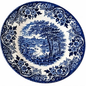May include: A round ceramic plate featuring a blue and white transferware pattern. The center depicts a landscape with a person fishing by a river, trees, and distant buildings. The border is decorated with intricate floral and scroll motifs.