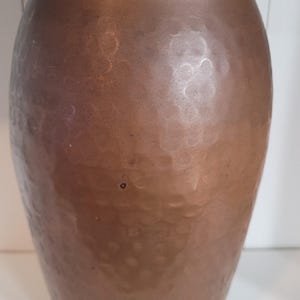 May include: A tall, cylindrical copper vase featuring a distinct hammered texture across its entire surface. The vessel has a wide, flared rim at the top and tapers slightly toward the base, showcasing a warm, metallic brown finish throughout.
