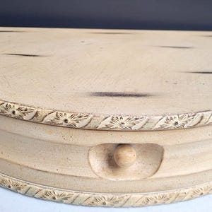 May include: A half-moon shaped, light beige wooden box with a decorative floral border. The box has a small, round knob and a drawer. The top has dark streaks, and the box appears to be aged.