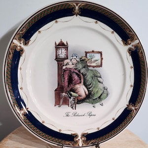 May include: A white decorative plate with a blue and gold border. The center features a detailed illustration of a couple embracing near a grandfather clock. The text "The Pickwick Papers" is below the illustration.