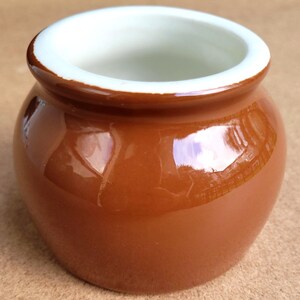 May include: A small, round ceramic pot with a glossy, solid brown exterior and a smooth, white interior rim. The vessel features a wide, open top and a curved, bulbous body, showcasing a reflective surface that highlights its simple, functional form.