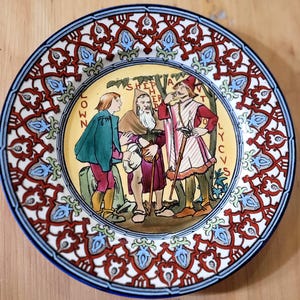 May include: A decorative ceramic plate featuring a central illustration of three figures in historical attire with the text "SHEPHERD" and "CLOWN" visible. The border displays a repeating red, blue, and white geometric pattern with floral motifs.