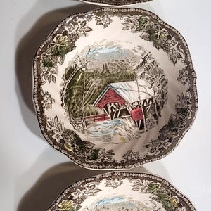May include: Three ceramic bowls featuring a central transferware illustration of a red covered bridge in a winter landscape. The scalloped rims are decorated with a detailed brown and green grape vine pattern on a cream-colored background.
