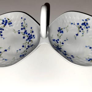May include: White serving dish with two oval compartments and a handle. The dish is decorated with blue floral patterns and green stems. The dish is made of porcelain.