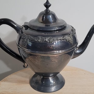 May include: A silver-colored teapot with a black handle and spout. The teapot has a decorative floral design around the middle and a lid with a finial. The teapot sits on a pedestal base.