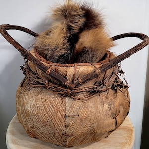 May include: A handcrafted, rustic basket with a rounded body and two arched handles. The basket is made of natural materials, with a textured, light brown exterior and woven details. It contains a fluffy, brown and black fur element.