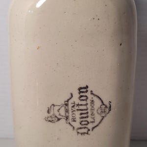 May include: Cream-colored ceramic bottle with a textured, light brown stopper. The bottle has a rounded shape and a black and white emblem that reads "Royal Doulton London."