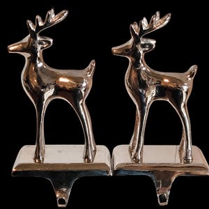 May include: A pair of shiny silver-toned metal stocking holders, each featuring a standing deer figure with antlers. The deer are mounted on rectangular bases with integrated hooks extending downward for hanging items.