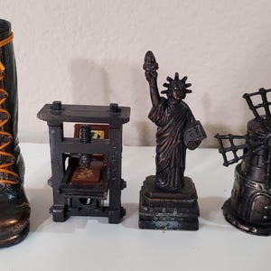 May include: A collection of miniature bronze-colored figurines. Includes a boot with orange laces, a printing press, the Statue of Liberty, and a windmill. The figurines are displayed on a white surface.