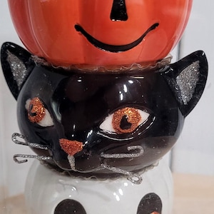 May include: A stacked Halloween decoration featuring a smiling orange pumpkin, a black cat, and a white skull. Each figure has glittery eyes and decorative accents. The cat has silver whiskers.