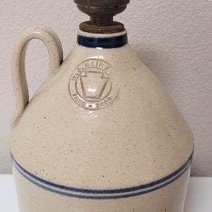 May include: A beige stoneware jug lamp with a speckled finish and two horizontal blue stripes. The jug features a stamped H.J. Heinz Co. logo with a keystone shape and the text 1869-1909. A brass-colored metal lamp socket sits on the top opening.
