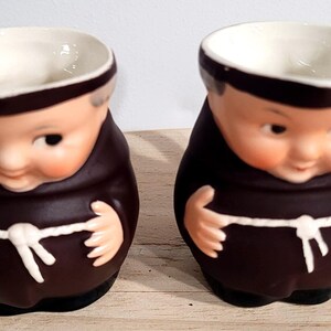 May include: Two ceramic monk-shaped containers. The containers are brown with white accents and feature a smiling face. The interior is white. The containers are likely used for holding small items or as decorative pieces.