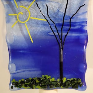 May include: A decorative night light featuring a fused glass design. The artwork depicts a yellow sun, a black tree, and green and black foliage against a blue and white sky. The night light has a white base.
