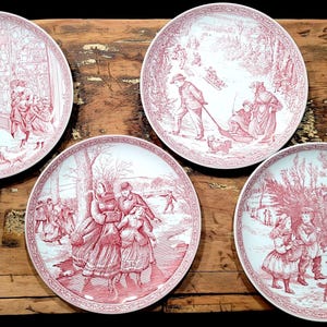 May include: Four round ceramic plates featuring detailed red and white transferware illustrations of winter scenes. Each plate depicts different outdoor activities including people walking, sledding, and carrying a Christmas tree in snowy landscapes.