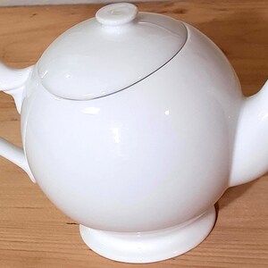 May include: A glossy white ceramic teapot with a spherical body, a curved handle, a short spout, and a matching lid with a small knob. The teapot features a smooth, rounded design and sits on a circular pedestal base, presenting a clean aesthetic.