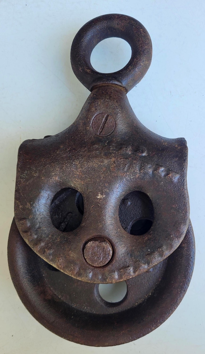 Puede incluir: Polea met&aacute;lica oxidada y antigua, con un lazo superior circular y una rueda. La polea tiene un aspecto marr&oacute;n oscuro y desgastado, con signos visibles de edad y uso. El dise&ntilde;o incluye dos aberturas circulares y una rueda central.
