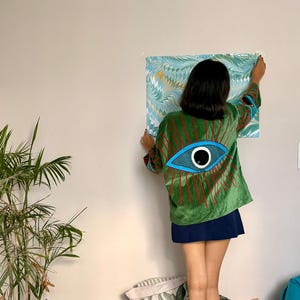 May include: A green velvet jacket with a large blue eye design on the back, featuring red vertical lines radiating outward. The jacket is paired with a navy blue skirt. A piece of art with blue and white patterns is being hung on the wall.