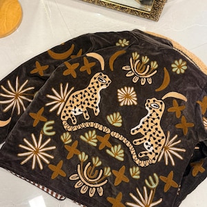 Puede incluir: Una chaqueta de terciopelo marr&oacute;n oscuro con bordados intrincados de dos leopardos manchados, lunas crecientes y varios motivos florales y estelares en tonos bronceado, crema y verde claro. La chaqueta tiene un cuello estructurado.