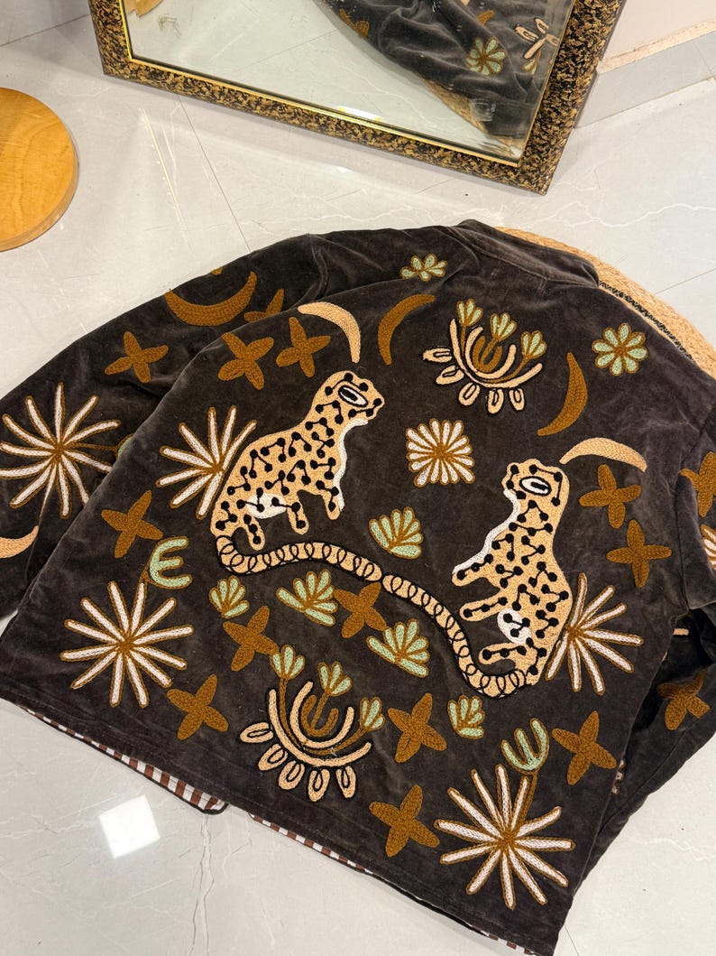Puede incluir: Una chaqueta de terciopelo marr&oacute;n oscuro con bordados intrincados de dos leopardos manchados, lunas crecientes y varios motivos florales y estelares en tonos bronceado, crema y verde claro. La chaqueta tiene un cuello estructurado.