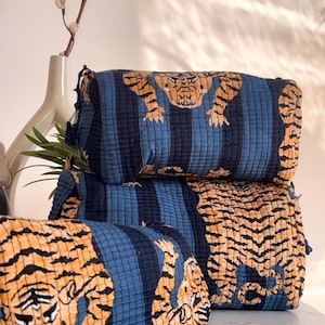 May include: Three rectangular, quilted cosmetic bags with a tiger print. The bags are navy blue with vertical stripes and orange tiger illustrations. The bags are stacked on top of each other.