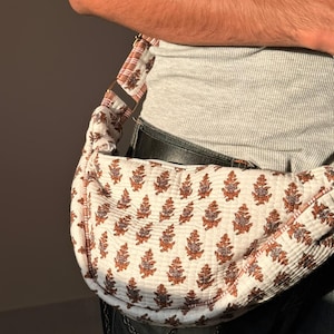 Op de afbeelding: Een witte en bruine heuptas met bloemenpatroon. De tas wordt over het lichaam gedragen en heeft een bijpassende riem. De tas is gemaakt van een zachte, gewatteerde stof.