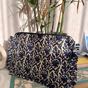 May include: A navy and cream floral patterned quilted cosmetic bag. The bag features a zip closure and a tassel detail. The rectangular bag is likely made from cotton or a similar material. The bag is a stylish accessory.