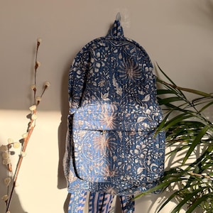 Puede incluir: Una mochila azul y blanca con estampado floral y diseño acolchado. La mochila tiene un bolsillo frontal y correas ajustables. La bolsa está colgada en una pared. Las medidas no son visibles.