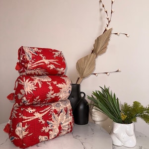 Może przedstawiać: Trzy czerwone poduszki cylindryczne z białym i niebieskim wzorem kwiatowym są ułożone w stos. Każda poduszka ma czerwoną frędzlę na każdym końcu. Na stole z marmurowym blatem, z dekoracyjnymi wazonami i roślinami w tle.