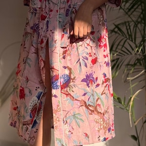 Może przedstawiać: Jasnoróżowy szlafrok z kwiatowym i ptasim wzorem. Szlafrok posiada białe wykończenie i rozcięcie z boku. Wzór zawiera elementy czerwone, niebieskie, fioletowe i zielone. Szlafrok jest do kolan i ma długie rękawy.