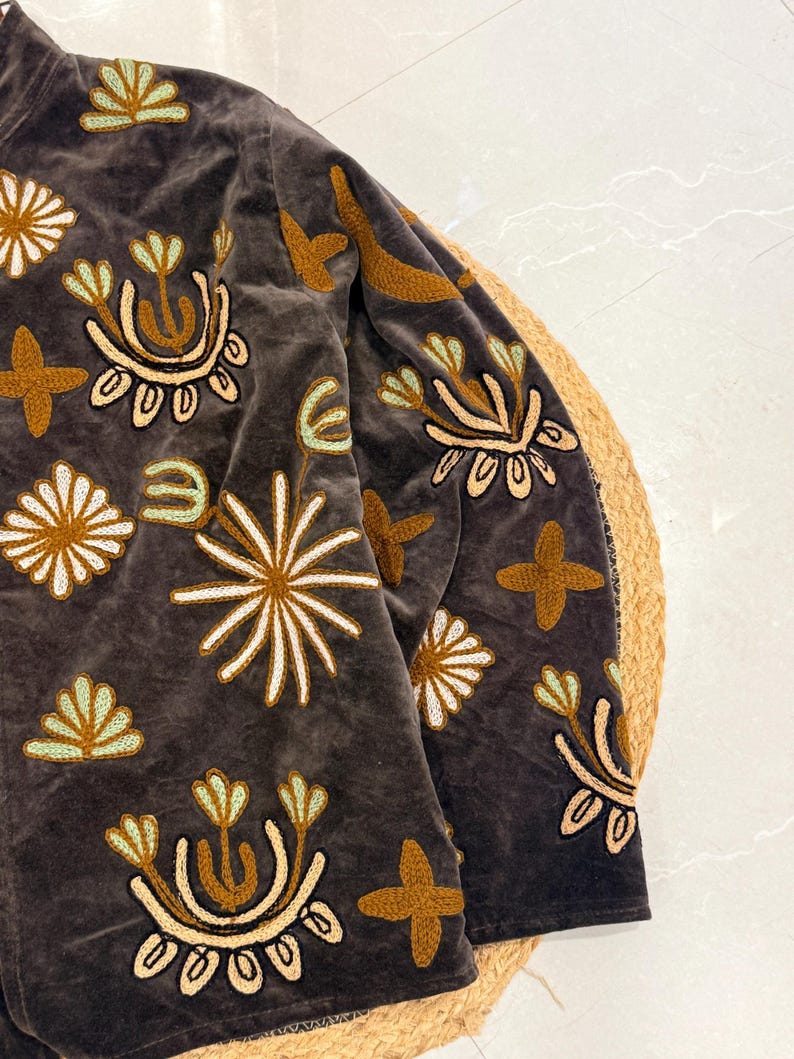 Puede incluir: Una chaqueta de terciopelo gris oscuro con bordados intrincados en tonos amarillo mostaza, crema y verde menta claro. El dise&ntilde;o consiste en varios motivos florales y en forma de estrella repartidos por la parte delantera y la manga larga.