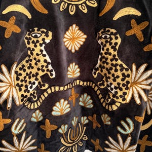 Puede incluir: Un tejido de terciopelo gris oscuro con un intrincado bordado de dos leopardos moteados frente a frente. El dise&ntilde;o incluye motivos florales, lunas crecientes y formas de cruz en amarillo dorado y azul claro dispuestos sim&eacute;tricamente.