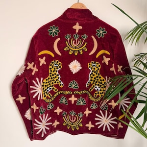 Puede incluir: Una chaqueta de terciopelo rojo oscuro con bordados intrincados de dos leopardos manchados en amarillo y negro, lunas crecientes y varios motivos florales y en forma de estrella en blanco, verde y amarillo en la espalda y las mangas.