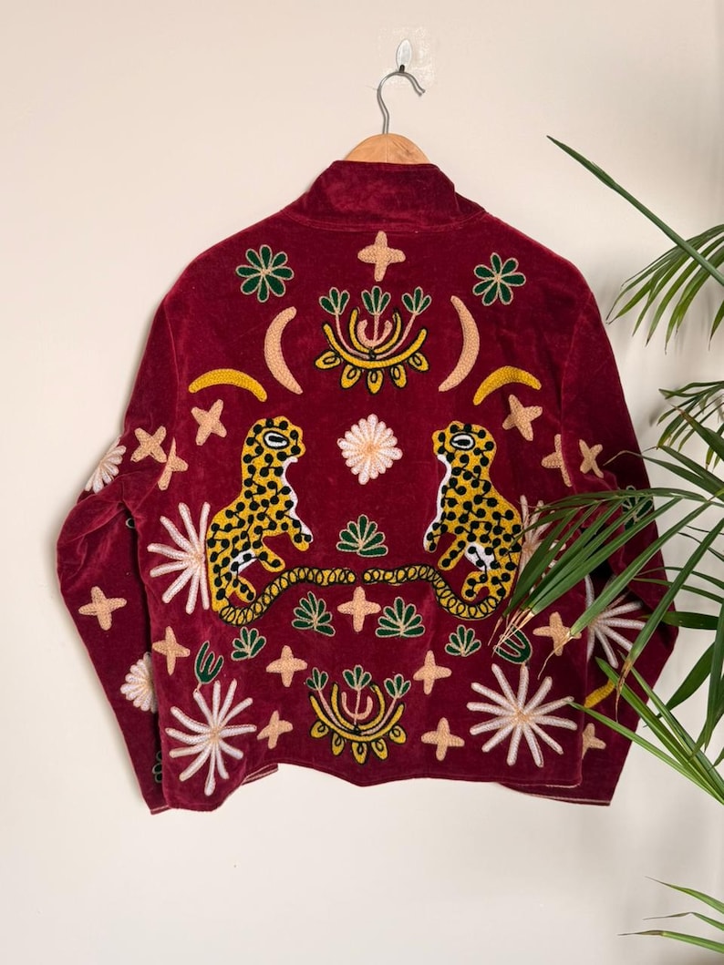 Puede incluir: Una chaqueta de terciopelo rojo oscuro con bordados intrincados de dos leopardos manchados en amarillo y negro, lunas crecientes y varios motivos florales y en forma de estrella en blanco, verde y amarillo en la espalda y las mangas.