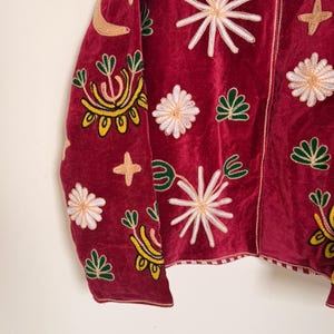 Puede incluir: Una chaqueta de terciopelo rojo oscuro con bordados intrincados de estrellas blancas, motivos florales verdes y lunas crecientes de color tostado. La prenda tiene un cierre de cremallera frontal y un borde a rayas en el dobladillo inferior.