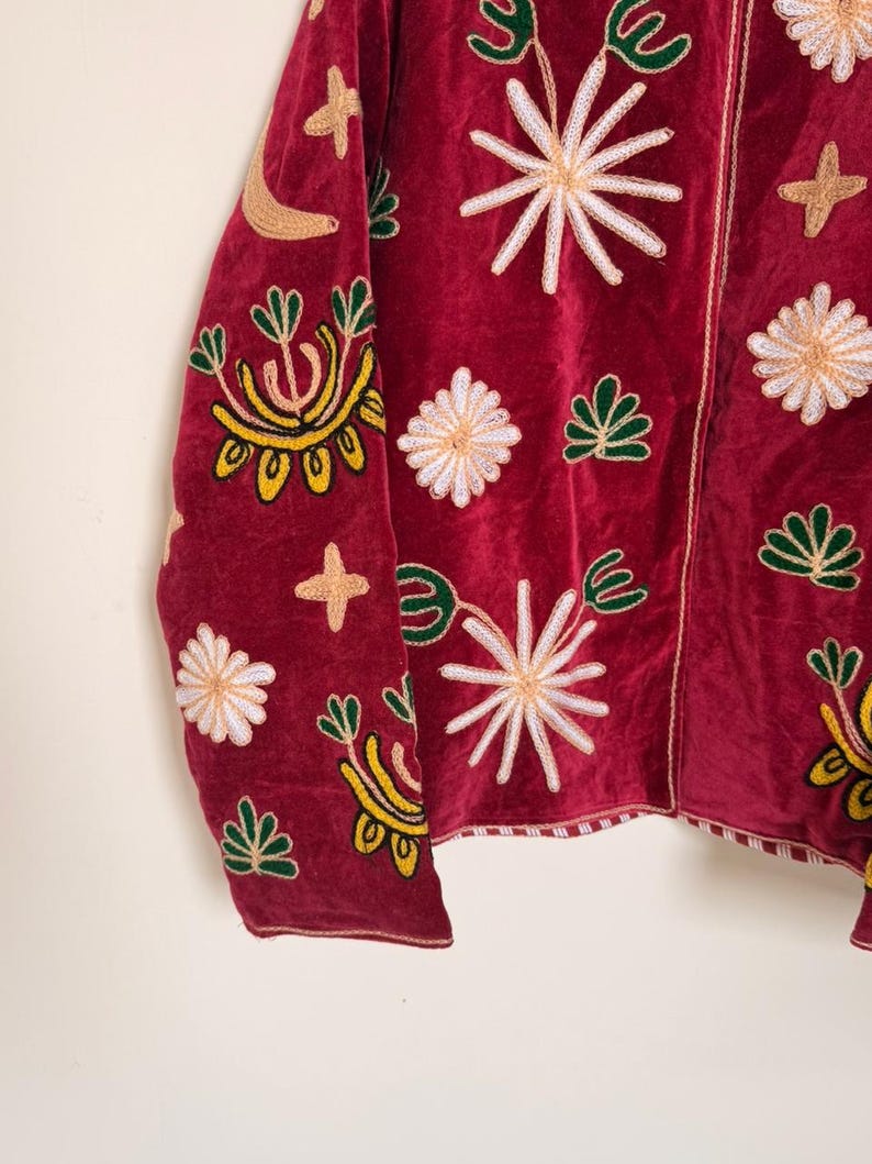 Puede incluir: Una chaqueta de terciopelo rojo oscuro con bordados intrincados de estrellas blancas, motivos florales verdes y lunas crecientes de color tostado. La prenda tiene un cierre de cremallera frontal y un borde a rayas en el dobladillo inferior.
