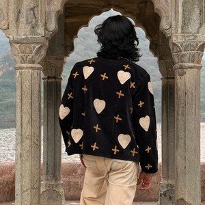 Puede incluir: Una chaqueta negra con corazones bordados en crema y cruces doradas. La chaqueta se lleva con pantalones color beige. La persona está de pie frente a una estructura de piedra arqueada, con un lago y montañas al fondo.