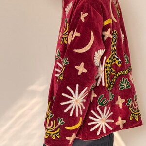 Puede incluir: Una chaqueta de terciopelo rojo intenso con bordados vibrantes y multicolores de varios motivos celestiales y animales, incluyendo estrellas, lunas crecientes y un leopardo. La chaqueta tiene un cuello alto y mangas largas con bordados.