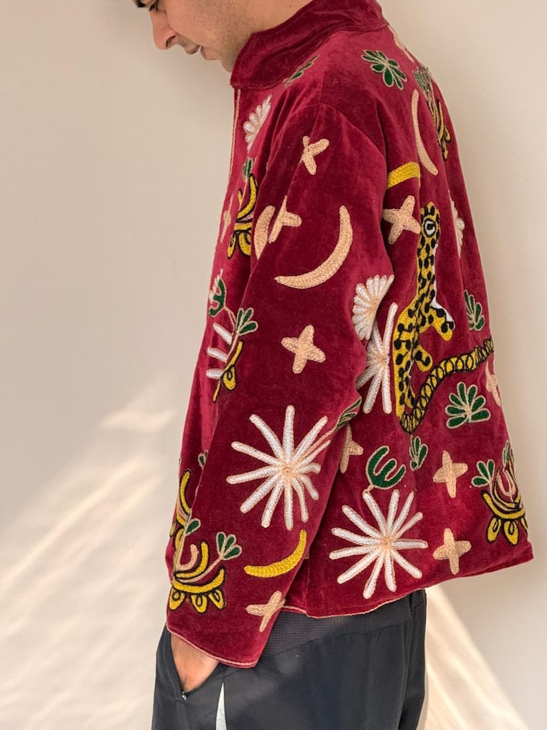 Puede incluir: Una chaqueta de terciopelo rojo intenso con bordados vibrantes y multicolores de varios motivos celestiales y animales, incluyendo estrellas, lunas crecientes y un leopardo. La chaqueta tiene un cuello alto y mangas largas con bordados.
