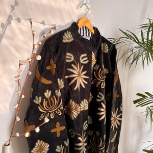 Puede incluir: Una chaqueta de terciopelo marr&oacute;n oscuro con intrincados bordados en tonos dorados, crema y azul claro. La chaqueta tiene cuello alto, mangas largas y un forro a rayas blancas y negras visible en el escote. El bordado representa motivos florales y abstractos.