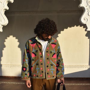 May include: An olive green jacket with vibrant floral embroidery in pink, blue, and black. The jacket features a button-up front and a white collar. The person is holding a black bag. The background is a white archway.