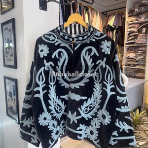 Peut inclure: Veste en velours noir avec broderie florale bleu clair. La veste a un col montant et une fermeture boutonnée. Le texte "Rubyballcloset" est visible. Elle est suspendue à un cintre en bois.