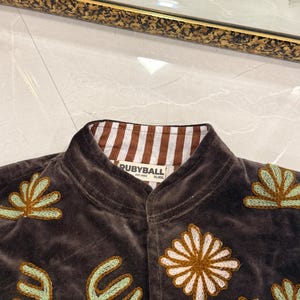 Puede incluir: Una chaqueta de terciopelo marr&oacute;n oscuro con cuello mao y forro interior a rayas marrones y blancas. La prenda est&aacute; decorada con motivos florales y bot&aacute;nicos bordados en tonos verde claro, blanco y marr&oacute;n. Una etiqueta muestra el texto RUBYBALL.