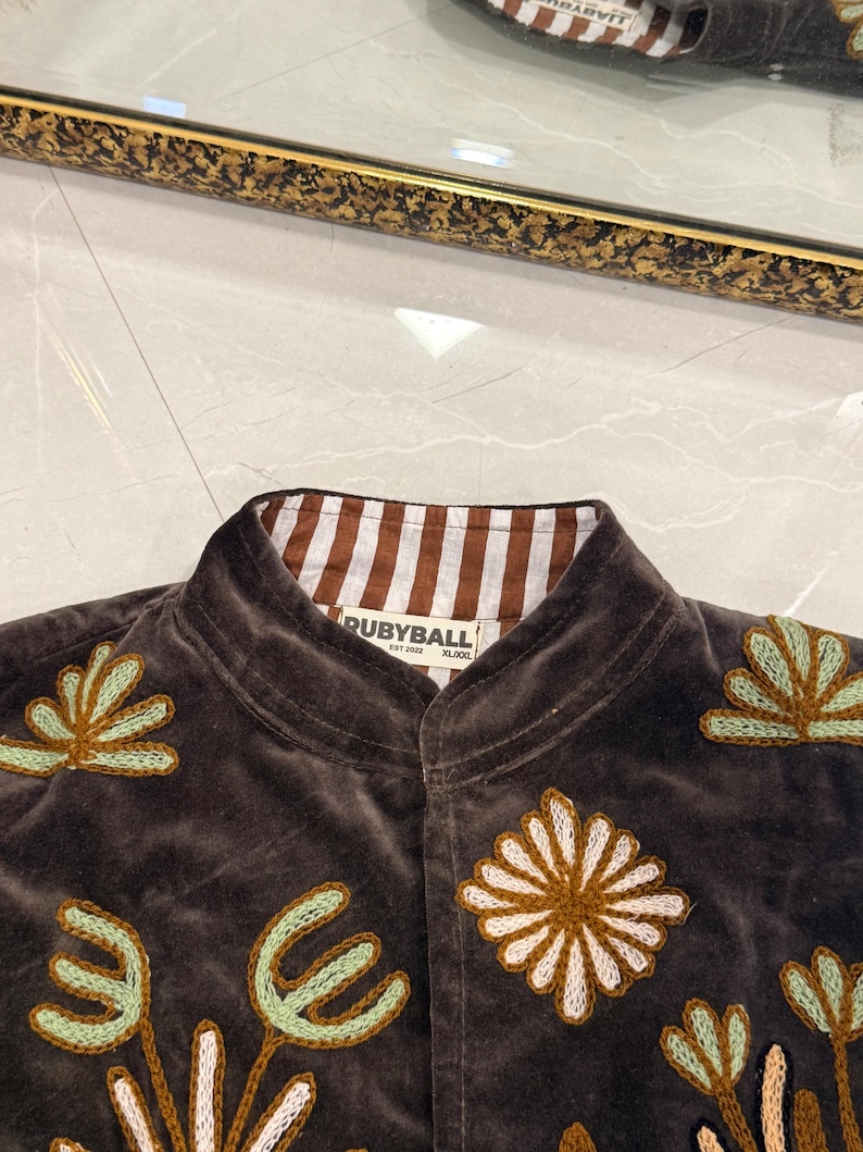 Puede incluir: Una chaqueta de terciopelo marr&oacute;n oscuro con cuello mao y forro interior a rayas marrones y blancas. La prenda est&aacute; decorada con motivos florales y bot&aacute;nicos bordados en tonos verde claro, blanco y marr&oacute;n. Una etiqueta muestra el texto RUBYBALL.